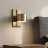 A modern bedroom with an aged brass Carmine LED Wall Sconce mounted on a textured beige wall beside a gray upholstered headboard and wood nightstand.
