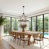A modern farmhouse dining room featuring a matte black Darlene 6 Light Chandelier 36" over a long wood table with woven rattan chairs and large black-framed windows.