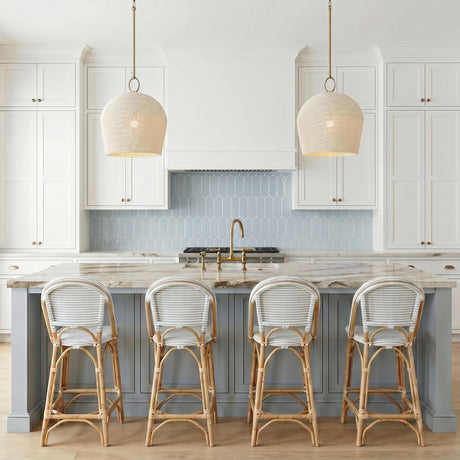 A coastal kitchen featuring two aged brass and ivory rattan pendants over a marble island with light blue cabinetry and a picket tile backsplash.