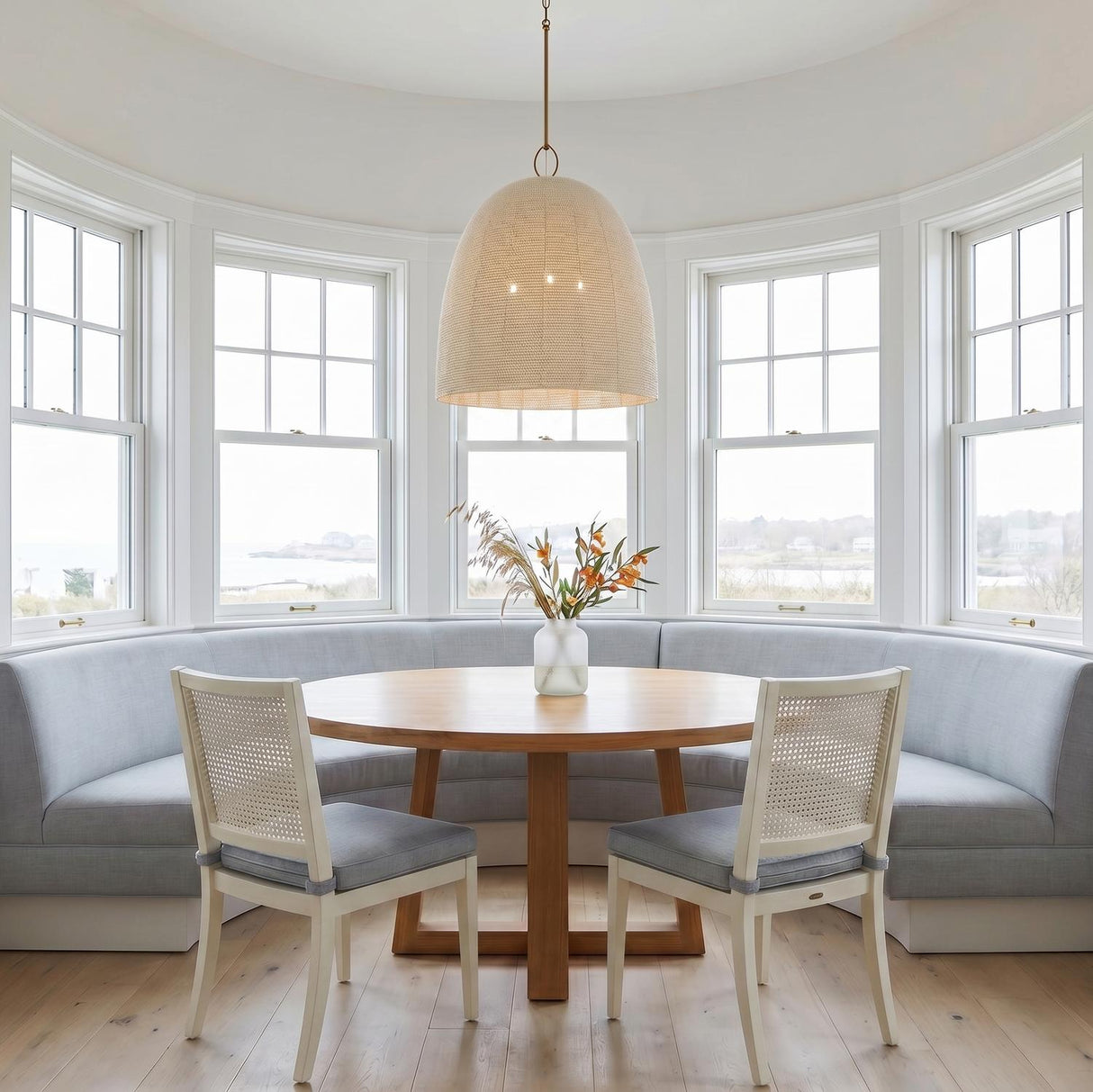 A coastal dining room featuring the Camiri Extra Large Pendant 25", Aged Brass and Ivory Beaded shade over a round wood table with a blue banquette and white cane chairs.