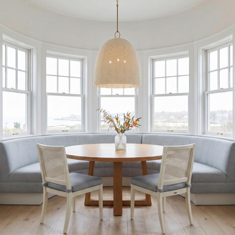 A coastal dining room featuring the Camiri Extra Large Pendant 25", Aged Brass and Ivory over a round wood table with a blue banquette and white cane chairs.