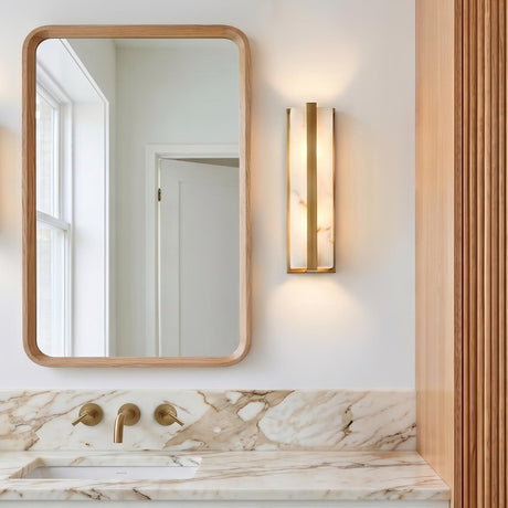 A modern bathroom with an Azura LED Wall Sconce 16", Aged Brass and Alabaster mounted beside a rounded oak mirror above a marble vanity counter.