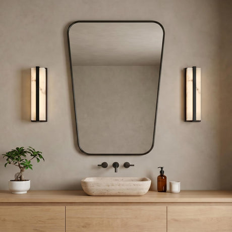 A minimalist bathroom with two matte black and alabaster wall sconces flanking a rounded rectangular mirror above a light wood vanity and stone vessel sink.