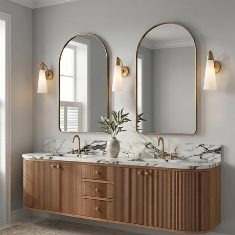A modern bathroom with three Ethel Wall Sconce fixtures in aged brass and opal glass flanking arched mirrors above a fluted wood vanity with marble countertops.