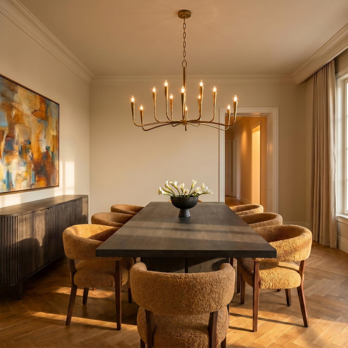 A contemporary dining room with the Myra 12 Light Chandelier 46" in artisan brass hanging over a dark wood table with textured mustard yellow chairs and herringbone floors.