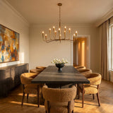 A contemporary dining room with the Myra 12 Light Chandelier 46" in artisan brass hanging over a dark wood table with textured mustard yellow chairs and herringbone floors.
