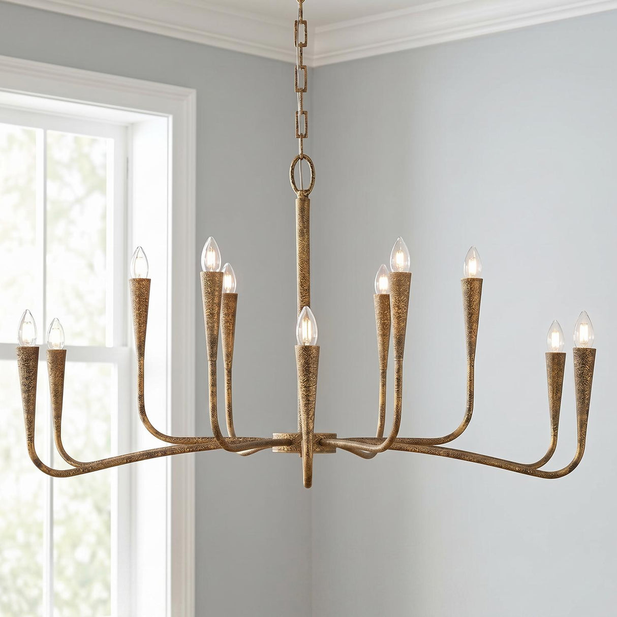 A transitional dining room with the Myra 12 Light Chandelier in a textured artisan brass finish centered beside a large window and light gray walls.