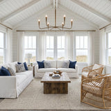 A coastal living room with the Myra 6 Light Chandelier 39", Artisan Brass centered under a vaulted shiplap ceiling above white slipcovered sofas and woven rattan chairs.