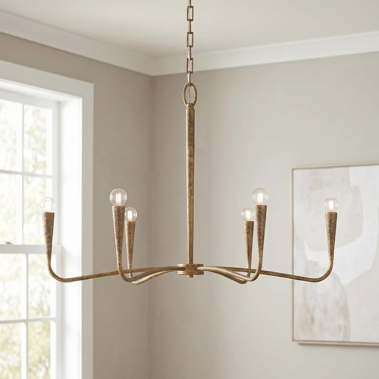 A transitional living room featuring the Myra 6 Light Chandelier 39", Artisan Brass in a textured finish suspended from the ceiling center near a large white-trimmed window.