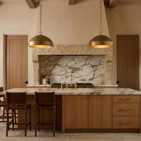 A Mediterranean kitchen with two artisan brass dome pendants over a white marble island featuring wood cabinetry, leather barstools, and rustic wood ceiling beams.