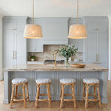 A coastal kitchen with two Malou Conical Pendants in natural rope and matte brass over a beige marble island with pale blue cabinetry and rattan stools.