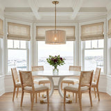 A coastal dining room with the Malou Drum Pendant in natural rope and matte brass over a white circular table and cane chairs under a coffered ceiling.
