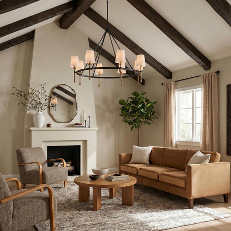A modern farmhouse living room with a matte black and hammered brass chandelier featuring white linen shades hanging from a vaulted beamed ceiling above a tan velvet sofa.