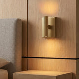 A modern bedroom with the Simeon Small LED Wall Sconce in Aged Brass mounted on light oak paneling beside a grey fabric headboard and matching nightstand.