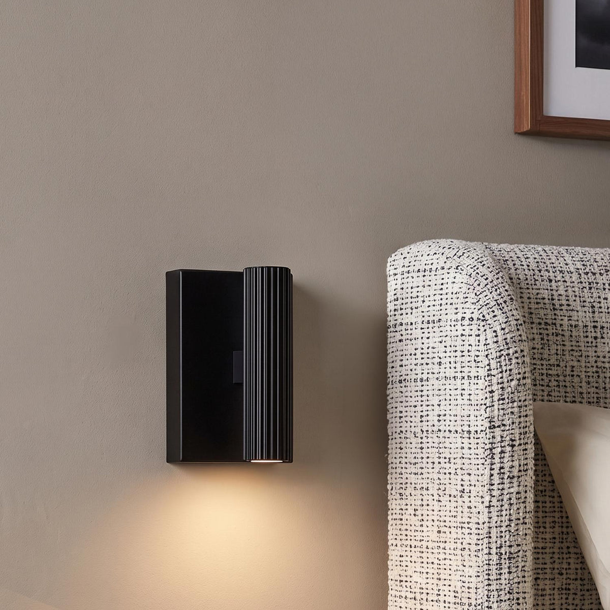 A modern bedroom with a matte black Simeon Small LED Wall Sconce mounted on a gray wall beside a textured woven fabric headboard.