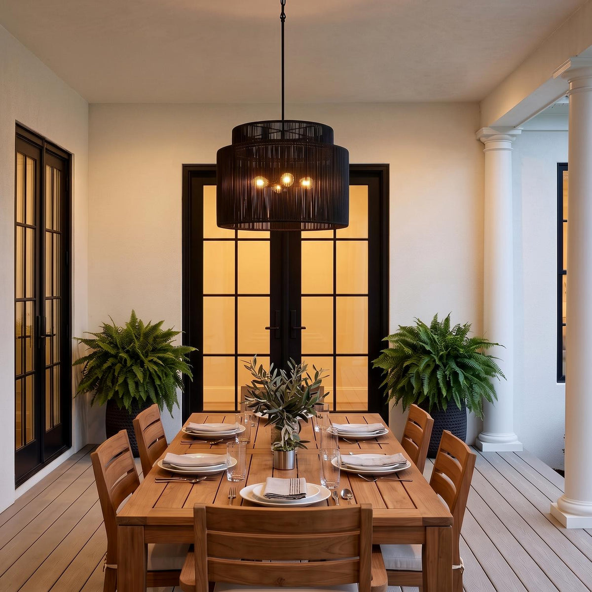 A transitional covered patio featuring the matte black Luma 4 Light Outdoor Pendant 29" glowing above a wood dining table against white stucco and black doors.