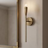 A modern bathroom with a brushed antique brass wall sconce mounted beside a gold-framed mirror on a textured cream wall under a standard ceiling.
