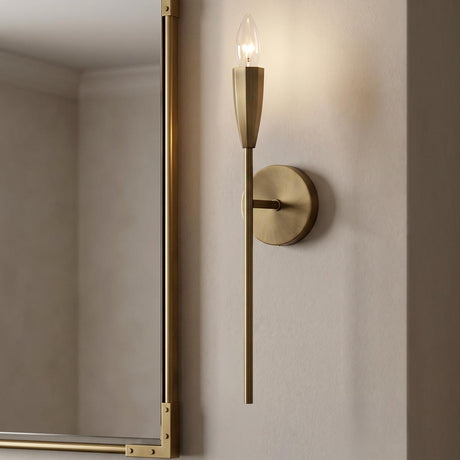 A modern bathroom with a brushed antique brass wall sconce mounted beside a gold-framed mirror on a textured cream wall under a standard ceiling.