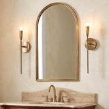 A transitional bathroom featuring two brushed antique brass wall sconces flanking an arched gold mirror above a tan marble vanity with matching brass fixtures.