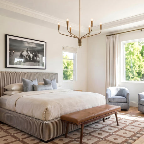 A transitional bedroom with the Rinaldo 6 Light Chandelier 35" in brushed antique brass over a striped bed, leather bench, and geometric patterned rug.