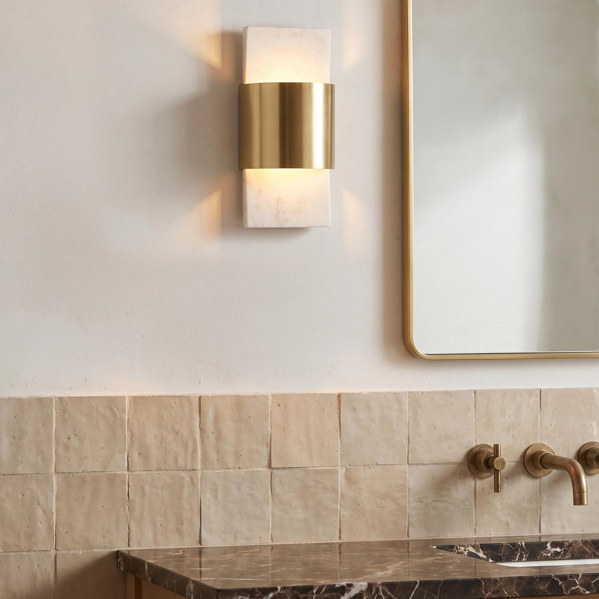 A transitional bathroom with the Solace LED Wall Sconce in white marble and matte brass mounted above a beige tile backsplash and dark marble vanity.