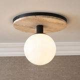 A modern foyer features a matte black Araceli flush mount with a white glass globe and travertine base mounted to the ceiling above textured tan grasscloth wallpaper.