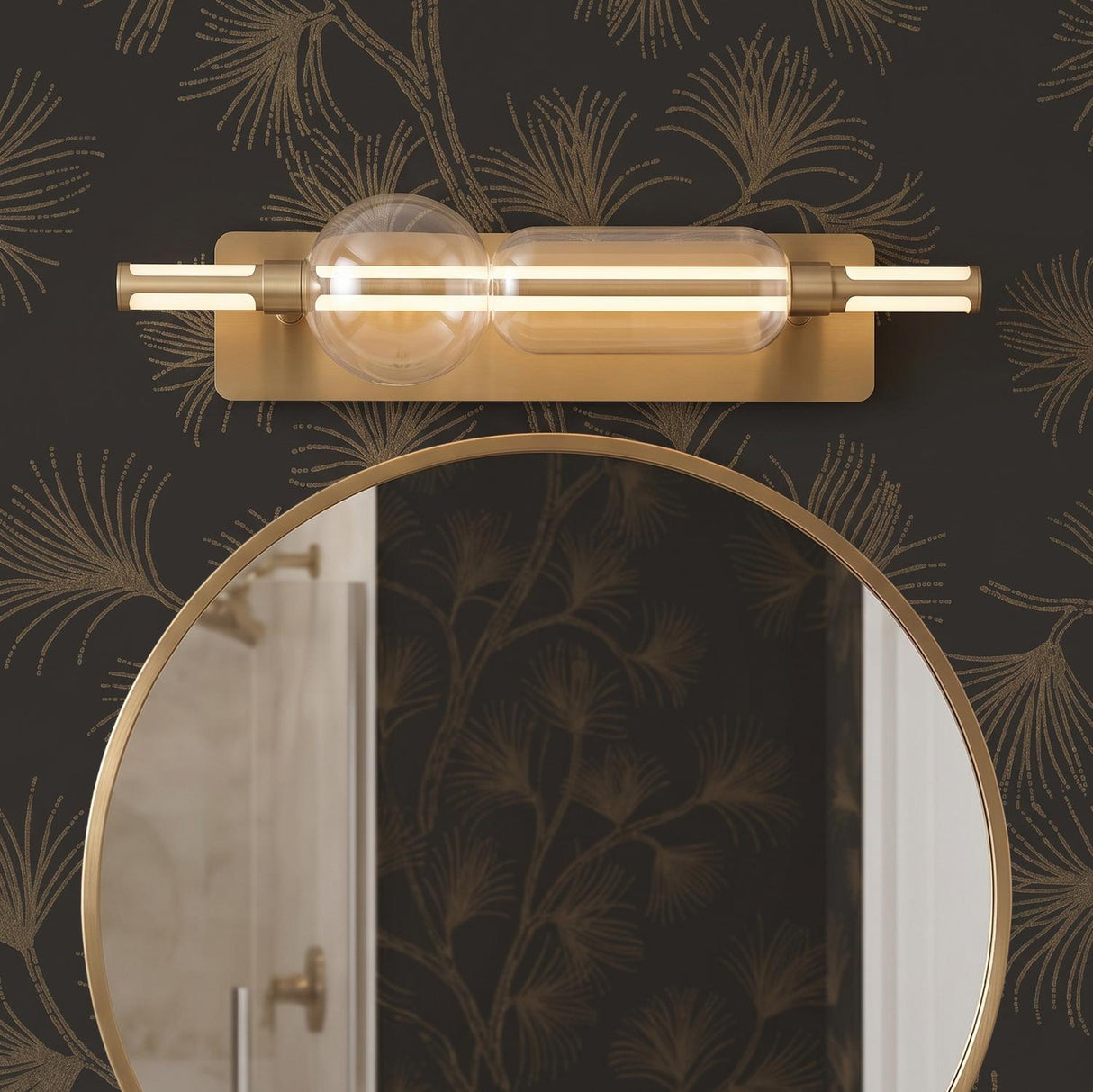 A modern bathroom features a brushed brass vanity bar with clear glass elements mounted above a round gold mirror on dark botanical patterned wallpaper.