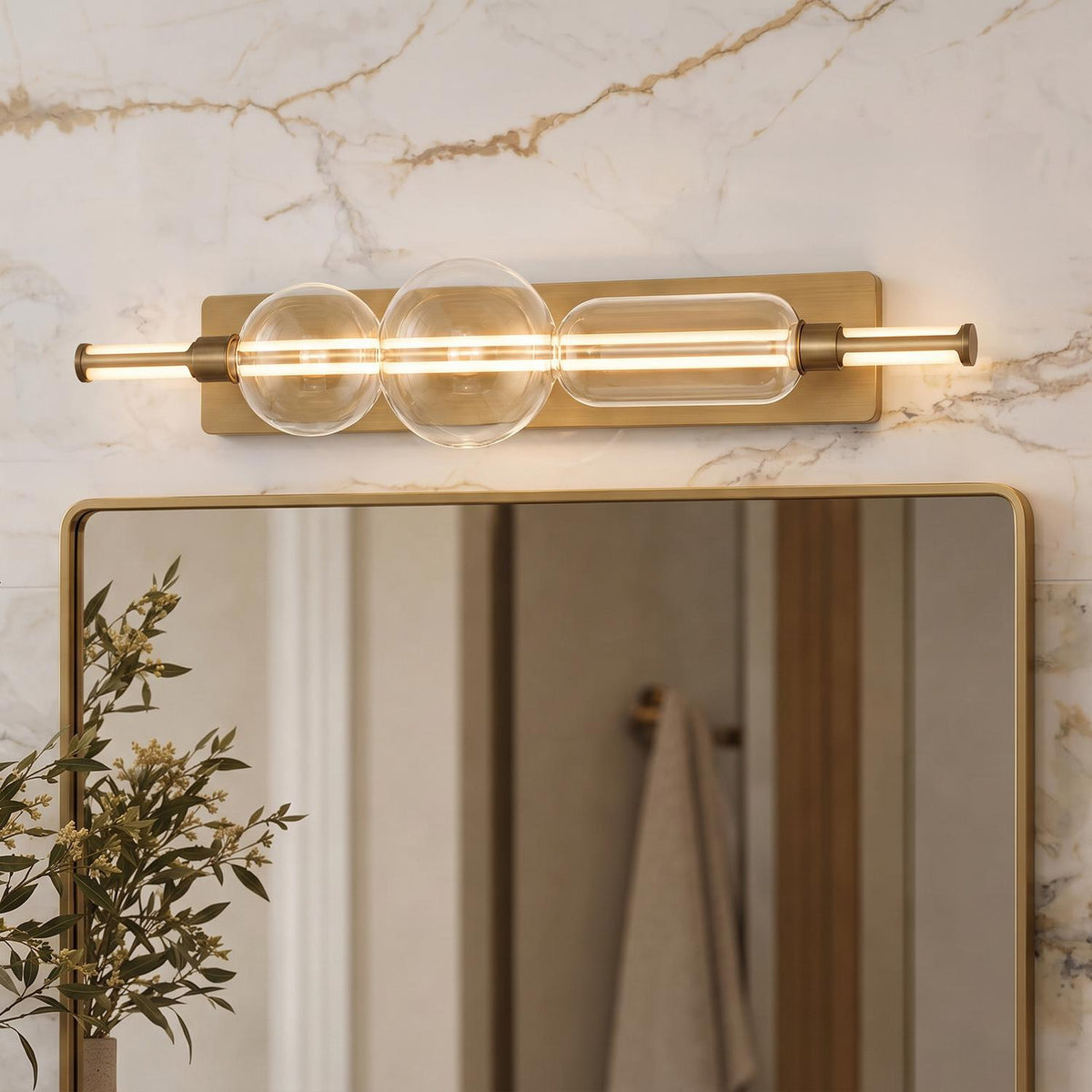 A modern bathroom features the Latimer LED Vanity Light in brass and clear glass mounted above a brass framed mirror on a white marble wall.
