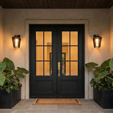 A Mediterranean home exterior with matte black and brass wall lanterns flanking a black double door on a stucco facade with large potted plants at dusk.