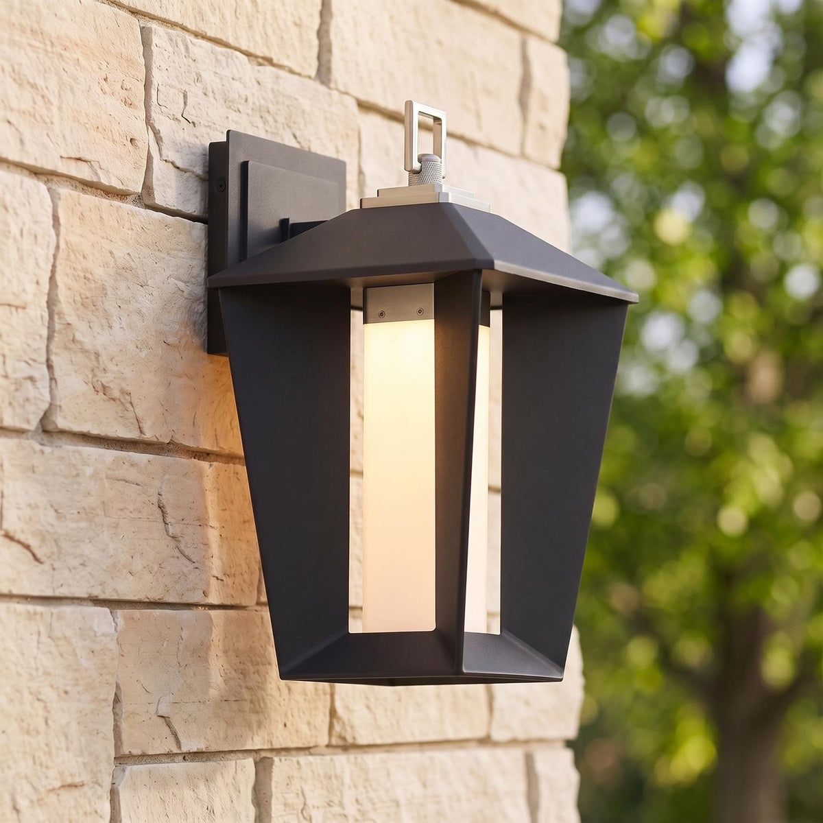 A modern home exterior with the Reynard LED Outdoor Wall Light in matte black and satin nickel glowing on stacked stone near lush green foliage at dusk.