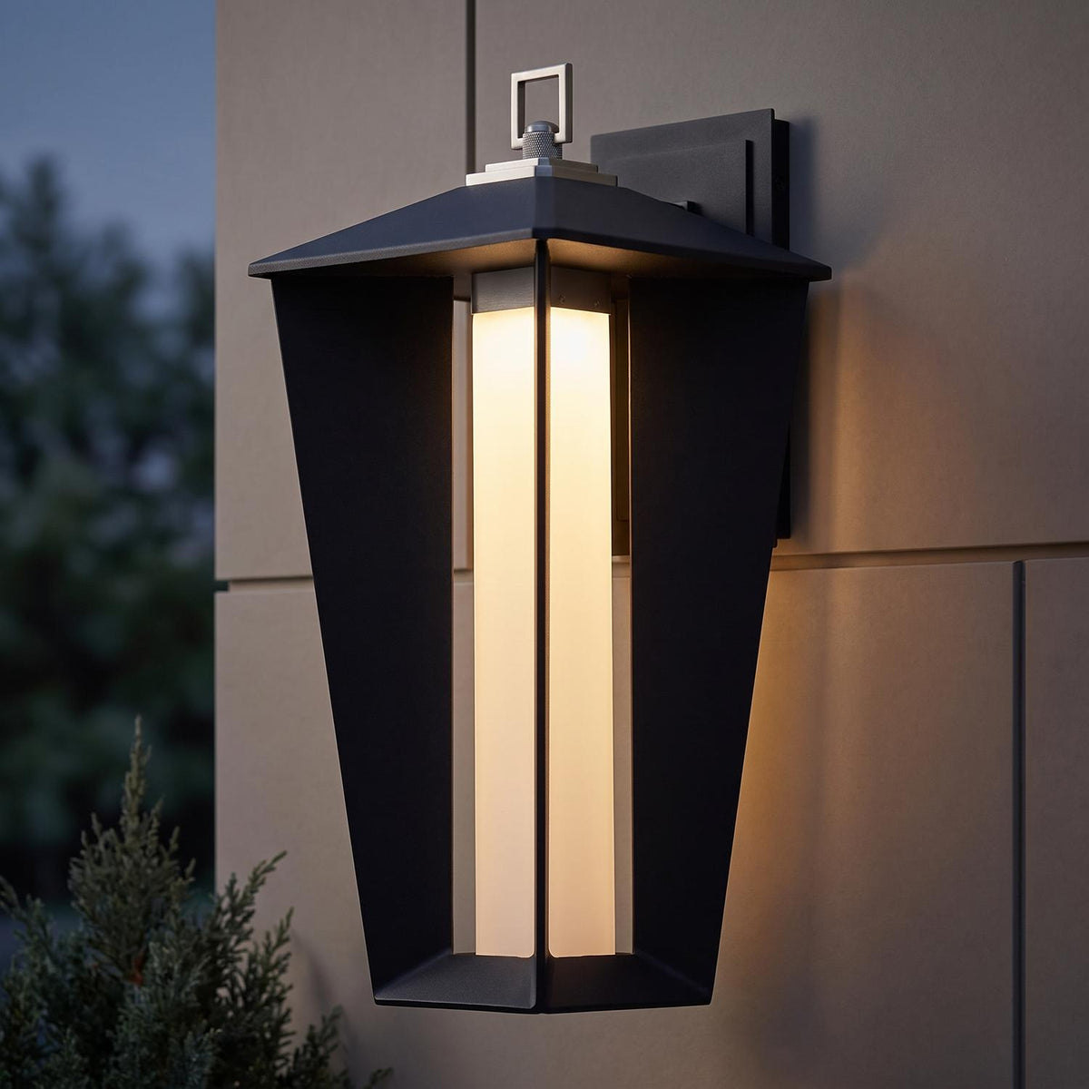 A modern home exterior featuring a Reynard LED Outdoor Wall Light in matte black and satin nickel mounted on a tan panel wall at twilight near evergreen landscaping.