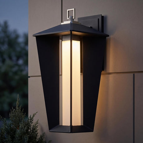 A modern home exterior featuring a Reynard LED Outdoor Wall Light in matte black and satin nickel mounted on a tan panel wall at twilight near evergreen landscaping.