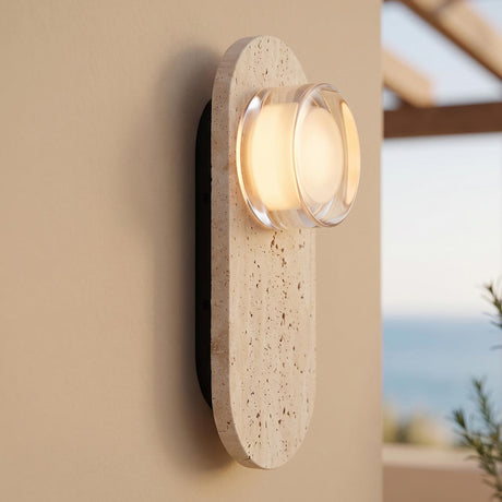 A modern coastal patio with a glowing wall sconce crafted from travertine and clear glass mounted on a beige stucco wall beside a wooden pergola.