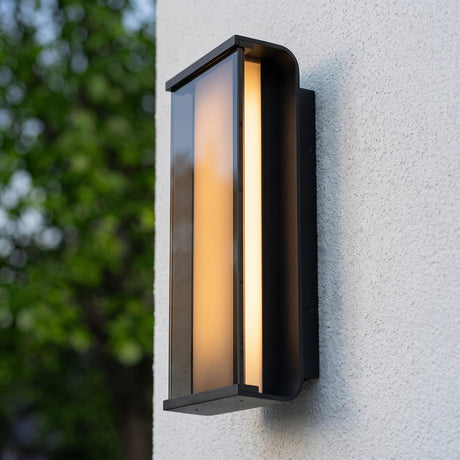 A modern home exterior with a glowing matte black Siran Outdoor LED Wall Sconce with smoke glass mounted on white stucco at dusk beside lush green trees.