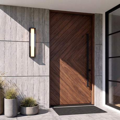 A modern entry with a glowing Siran Outdoor LED Wall Sconce in matte black mounted on a concrete wall beside a dark wood door and potted grasses.