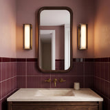 A mid-century bathroom with two Siran Medium LED Wall Sconces in antique brass and smoke glass flanking a walnut mirror above vertical burgundy wall tiles.