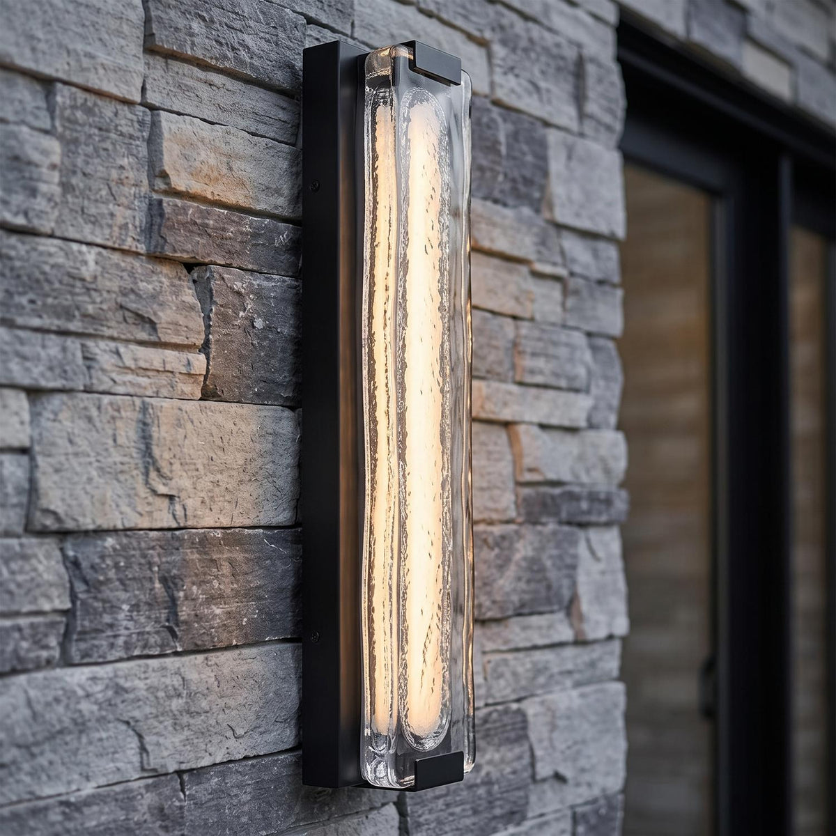 A modern home exterior featuring a glowing matte black wall sconce with textured glass mounted on a gray stacked stone wall beside a glass door at dusk.