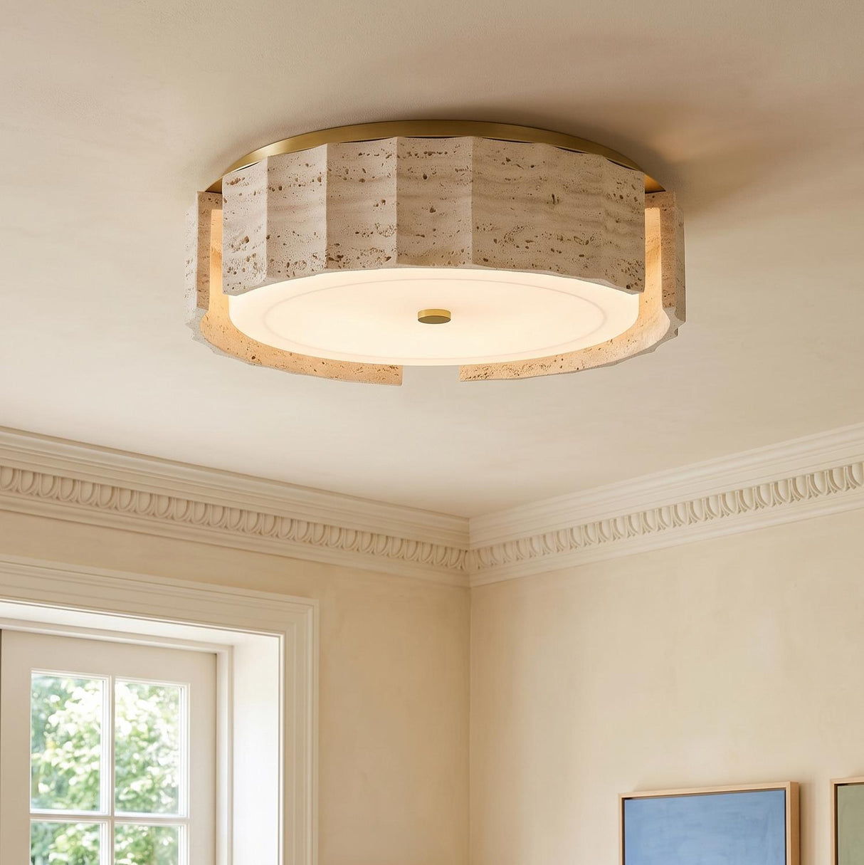 A contemporary living room with the Damari Medium LED Flush Mount in travertine and brass on the ceiling beside white crown molding and framed colorful artwork.