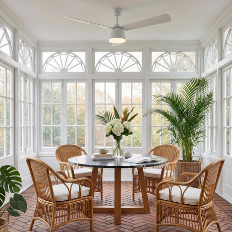 A coastal dining room featuring the Linnet LED Ceiling Fan 60-inch, Matte White over a black marble table and wicker chairs surrounded by white windows and brick flooring.