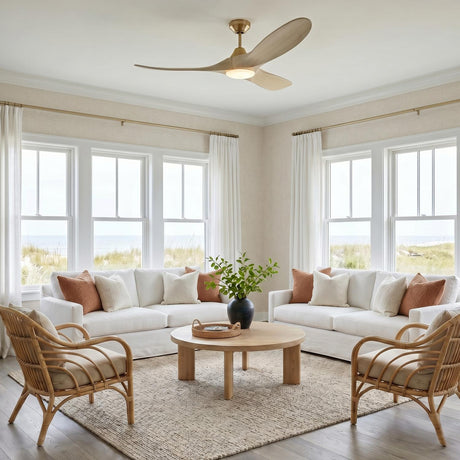 A coastal living room features the Egret LED Ceiling Fan 52", Whitewash Oak and Modern Brass centered above white slipcovered sofas, rattan armchairs, and beach views.