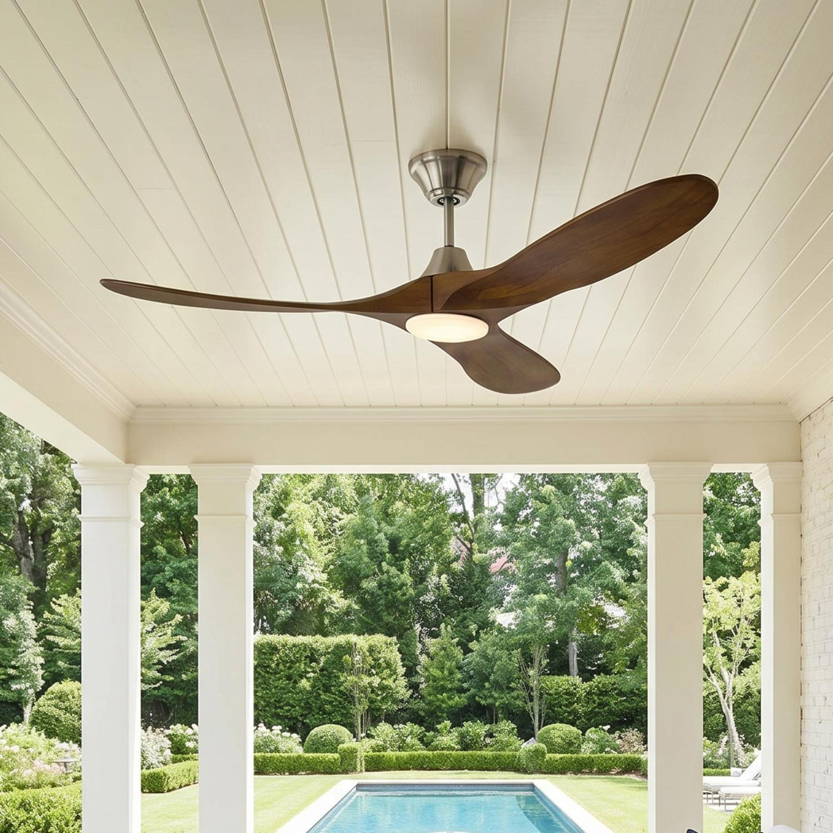 A traditional porch with white columns and the Egret LED Ceiling Fan 52", Walnut Wood and Brushed Steel mounted on a white ceiling overlooking a swimming pool.