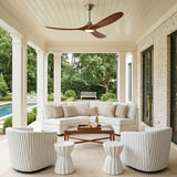 A modern farmhouse porch featuring the Egret LED Ceiling Fan 60" in koa wood and brushed steel mounted above a curved white sectional and white brick walls.