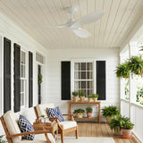 A modern farmhouse porch with white siding and black shutters featuring the matte white Heron Outdoor Smart Ceiling Fan 52 inch above wooden armchairs and potted ferns.