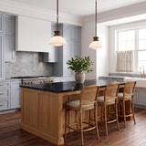 A transitional kitchen with two vintage bronze and milk glass pendants over a black marble island with blue-grey cabinetry and rattan bar stools.