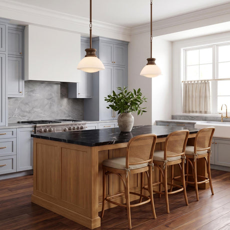 A transitional kitchen with two vintage bronze and milk glass pendants over a black marble island with blue-grey cabinetry and rattan bar stools.