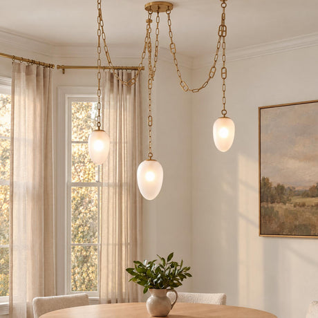 A transitional dining room with the Lockwood 3 Light Swag Chandelier, Vintage Brass and Frosted Glass draped above a round wooden table beside sheer curtains.