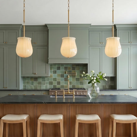 A transitional kitchen featuring three Cortland Medium Pendants in champagne glass and aged brass over a black soapstone island with reeded wood panels and muted green cabinetry.