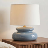 A modern bedroom featuring the Ellsworth Table Lamp 20", Blue Crackle Ceramic with a white linen shade on a fluted walnut nightstand beside a white wall.