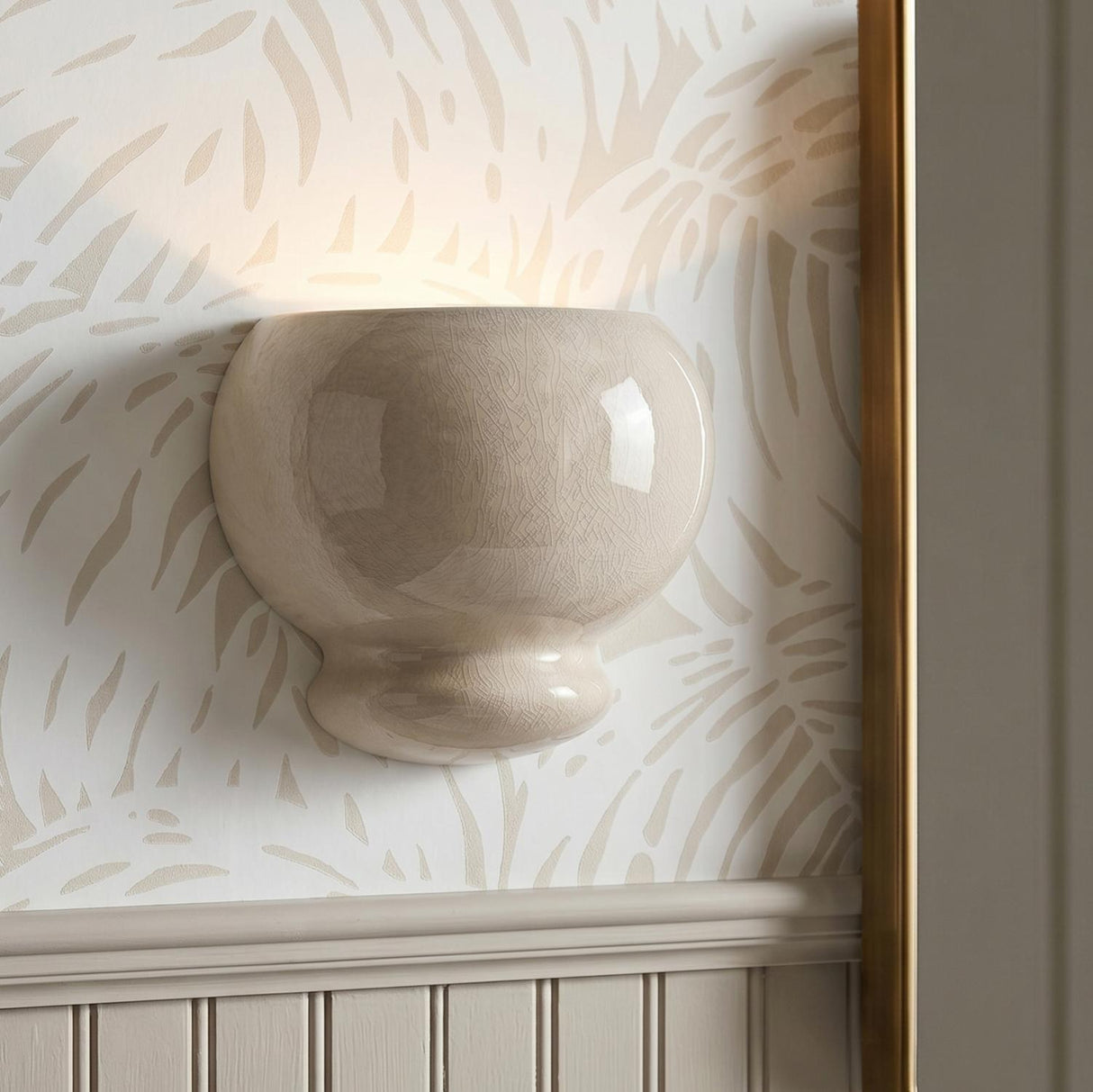 A transitional foyer with the Ellsworth Wall Sconce in Taupe Crackle Ceramic mounted on tropical leaf wallpaper above white wainscoting and a brass framed mirror.