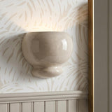 A transitional foyer with the Ellsworth Wall Sconce in Taupe Crackle Ceramic mounted on tropical leaf wallpaper above white wainscoting and a brass framed mirror.